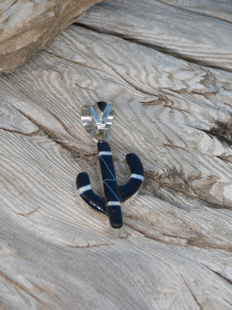 May include: A black cactus pendant with silver accents and a silver bail. The pendant has white and blue details. The pendant is displayed on a weathered wooden surface.