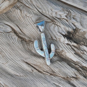 May include: A silver-tone cactus pendant with an opal inlay. The pendant has a detailed design with a textured surface. The cactus shape is a light blue color with white and pink accents. The pendant is displayed on a wooden surface.