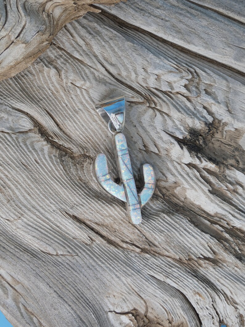May include: A silver-tone cactus pendant with an opal inlay. The pendant has a detailed design with a textured surface. The cactus shape is a light blue color with white and pink accents. The pendant is displayed on a wooden surface.