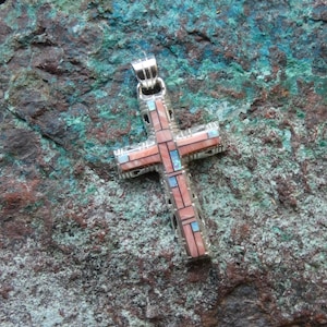 May include: A silver cross pendant featuring an intricate inlay of rectangular pink shell and small blue stone segments. The metal frame has a textured, geometric pattern along the edges and includes a silver bail at the top for hanging.