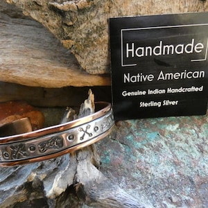 May include: A handmade Native American sterling silver cuff bracelet with an intricate design. The bracelet has a copper-colored interior and a silver exterior with etched patterns. A black card with white text reads "Handmade Native American Genuine Indian Handcrafted Sterling Silver."