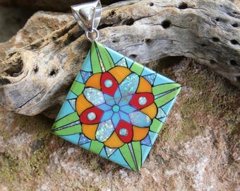 May include: A square pendant with a colorful geometric design featuring a blue, white, and red flower with a yellow center. The pendant is made of polymer clay and has a silver bail.
