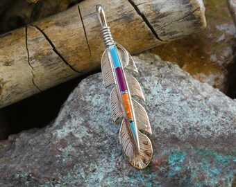 Könnte beinhalten: Silberfarbener Federanhänger mit eingelegten türkisen, lila und orangefarbenen Steinen. Der Anhänger hat detaillierte Federätzungen und eine silberne Öse für eine Halskette. Der Schmuck wird auf einem Stein- und Holzhintergrund präsentiert.