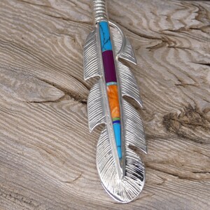 May include: A silver feather pendant with turquoise, orange, and purple inlay. The feather is detailed with etched lines and has a loop at the top for hanging.