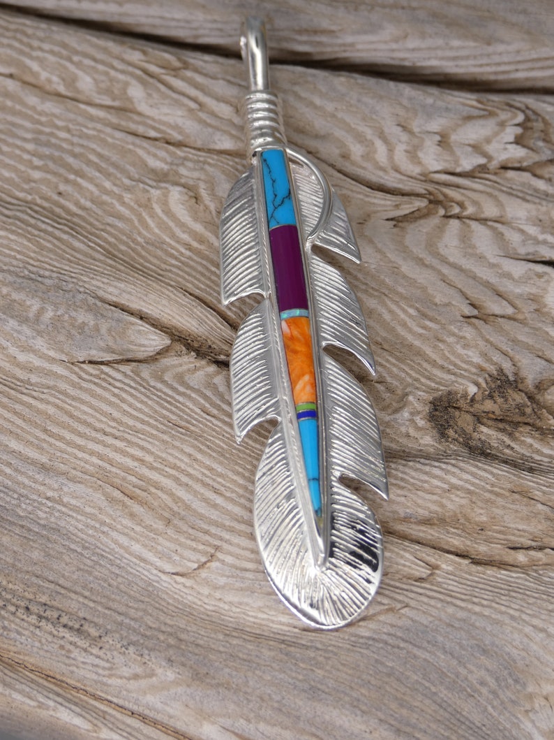 May include: A silver feather pendant with turquoise, orange, and purple inlay. The feather is detailed with etched lines and has a loop at the top for hanging.