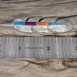 May include: A silver feather pendant with turquoise, purple, orange, and green inlay. The pendant is 2 inches long and is shown next to a ruler.