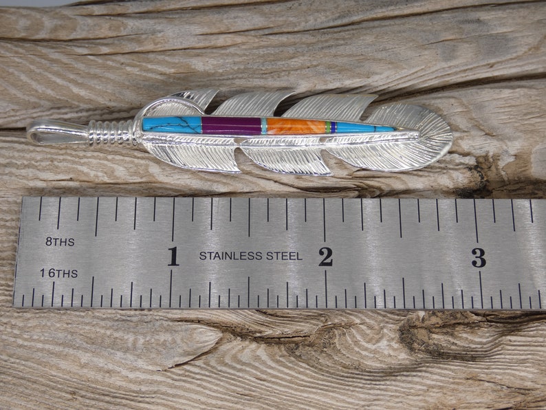 May include: A silver feather pendant with turquoise, purple, orange, and green inlay. The pendant is 2 inches long and is shown next to a ruler.