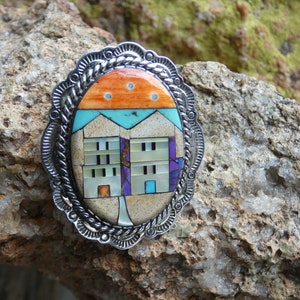 Puede incluir: Un colgante de plata e incrustaciones de turquesa con un diseño de mosaico multicolor de dos casas sobre un fondo blanco. El colgante está engastado en un marco ovalado de plata con un borde de cuerda.