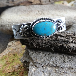 May include: A silver bracelet with a turquoise stone and decorative accents. The bracelet is on a wooden surface.