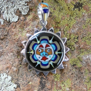 Puede incluir: Un colgante de plata con fondo negro y un diseño de cara de sol colorido. La cara del sol está rodeada por un diseño de rayos de sol de plata. El colgante está hecho de plata de ley e incrustado con turquesa, coral y otras piedras preciosas.