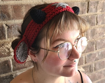 May include: A red crocheted headband with black and gray horns. The headband is worn by a person with a tattoo on their shoulder.