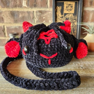 Puede incluir: Una bolsa de cordón de crochet negro con orejas rojas y un cordón rojo. La bolsa tiene una correa de crochet negro.