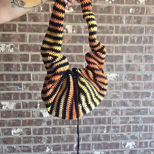 May include: A handmade crochet bag with a unique design. The bag features alternating stripes of black, orange, and yellow. The handles are long and curved, and the bag has a drawstring closure. The bag is held up against a brick wall.
