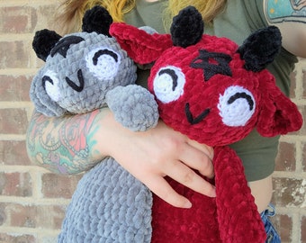 May include: Two crocheted plush toys, one gray and one red, with black horns and white eyes. The toys are being held by a person with a tattoo.