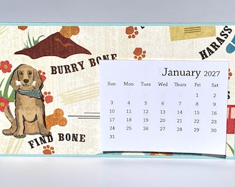 May include: A rectangular desk calendar for January 2027 featuring a light-colored background with illustrations of a brown dog holding a bone, red flowers, paw prints, and the text BURRY BONE and FIND BONE printed in a dark brown serif font.
