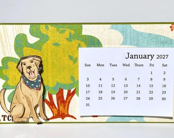 May include: A rectangular desk calendar for January 2027 featuring a yellow Labrador retriever wearing a blue bandana with white stars. The background displays a stylized tree with green leaves and a brown trunk, accented by orange paw prints.