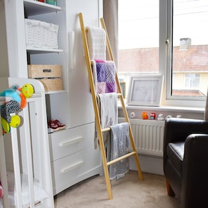 May include: A wooden ladder shelf with four shelves, holding blankets and towels in a nursery. The ladder shelf is made of natural wood and has a rustic look. The shelves are decorated with colorful blankets and towels. The ladder shelf is a great way to add storage and style to any room.