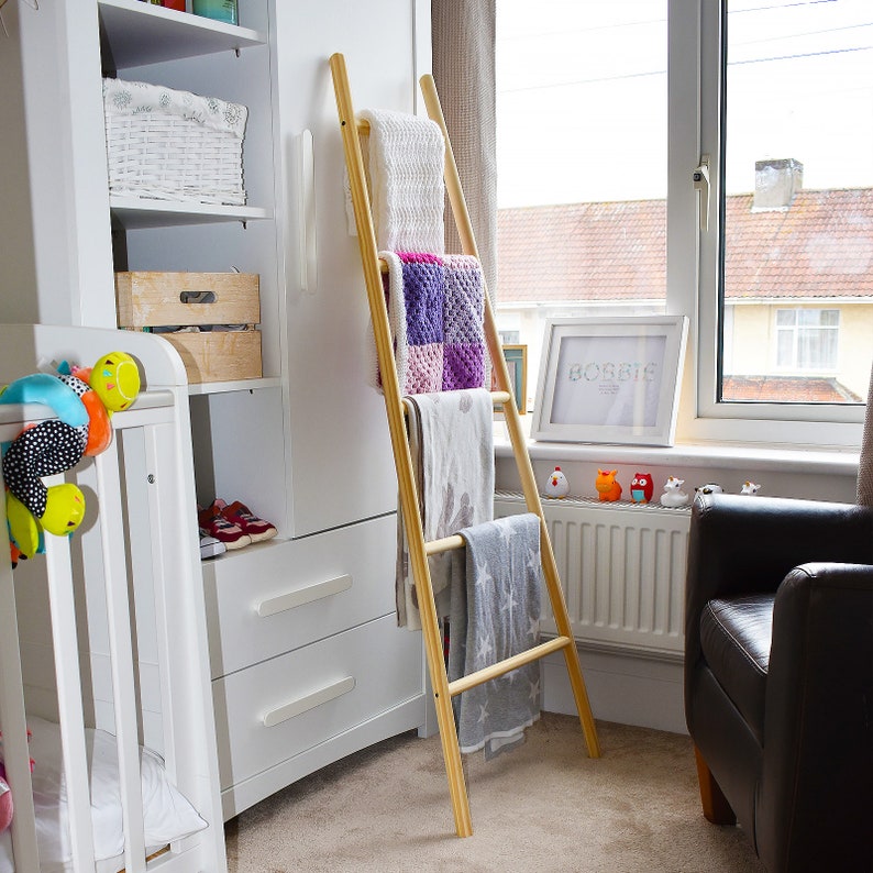 May include: A wooden ladder shelf with four shelves, holding blankets and towels in a nursery. The ladder shelf is made of natural wood and has a rustic look. The shelves are decorated with colorful blankets and towels. The ladder shelf is a great way to add storage and style to any room.