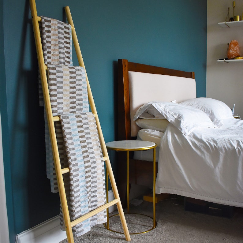 May include: A wooden ladder style towel rack with three towels folded on it. The towels are a light brown with white and blue stripes. The rack is in front of a teal wall and a white bed with a wooden headboard.
