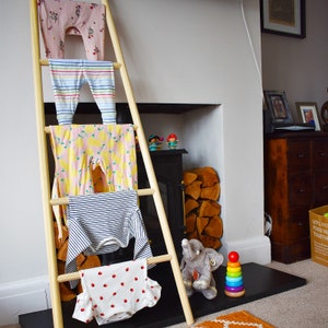May include: A wooden ladder with four rungs, each holding a different patterned baby garment. The garments are pink, blue, yellow, and white with red polka dots. The ladder is leaning against a wall with a fireplace and a wooden shelf above it. A stuffed elephant and a colorful stacking toy are on the floor in front of the ladder.