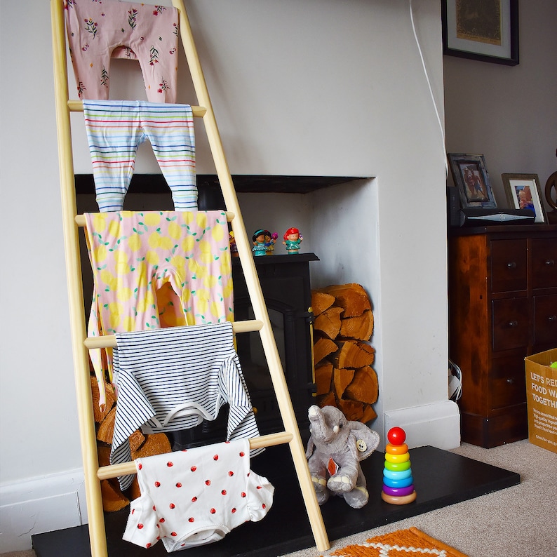 May include: A wooden ladder with four rungs, each holding a different patterned baby garment. The garments are pink, blue, yellow, and white with red polka dots. The ladder is leaning against a wall with a fireplace and a wooden shelf above it. A stuffed elephant and a colorful stacking toy are on the floor in front of the ladder.