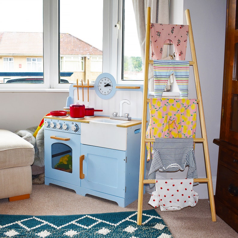 May include: A blue and white toy kitchen playset with a red stovetop and a white sink. A wooden ladder with clothes hanging on it is in the background.