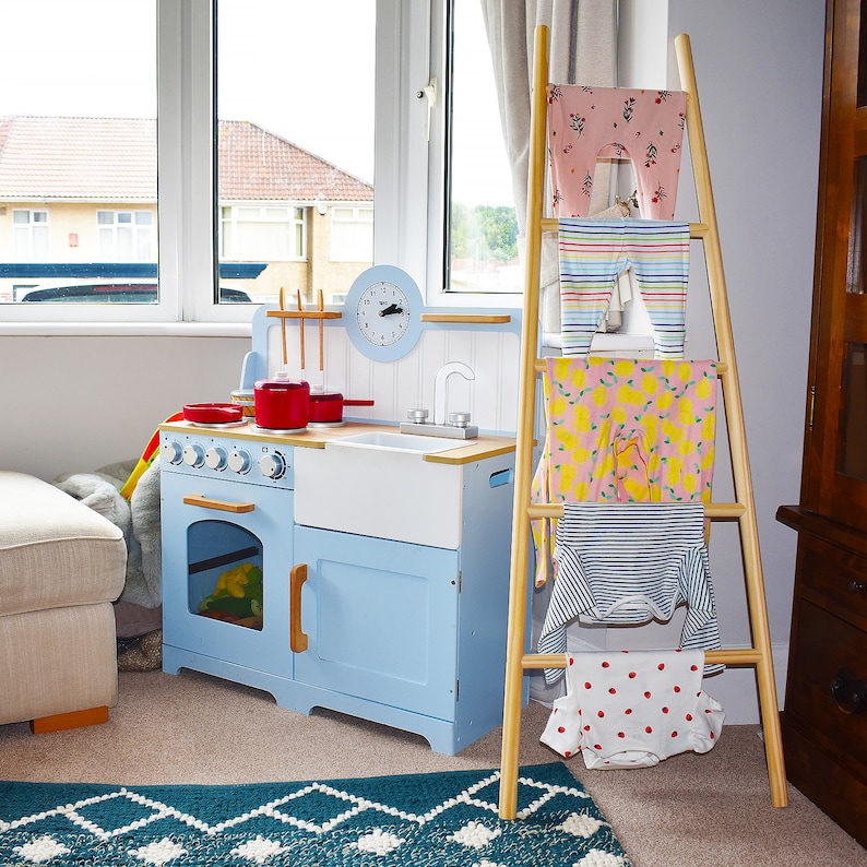 May include: A blue and white toy kitchen playset with a red stovetop and a white sink. A wooden ladder with clothes hanging on it is in the background.