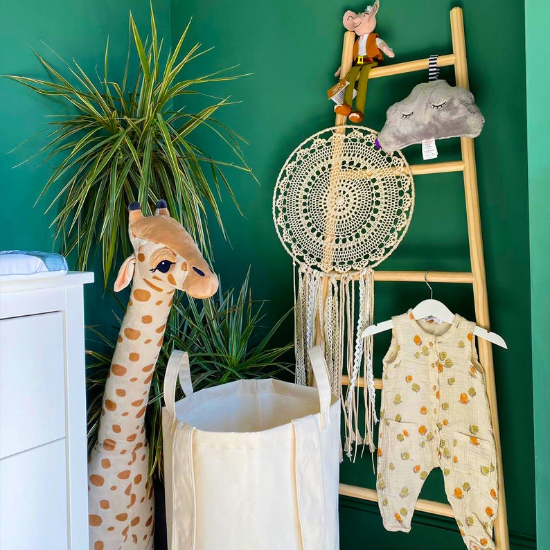 May include: A green wall with a wooden ladder, a white canvas laundry basket, a stuffed giraffe, a dream catcher, a stuffed animal, and a baby onesie hanging on the ladder.