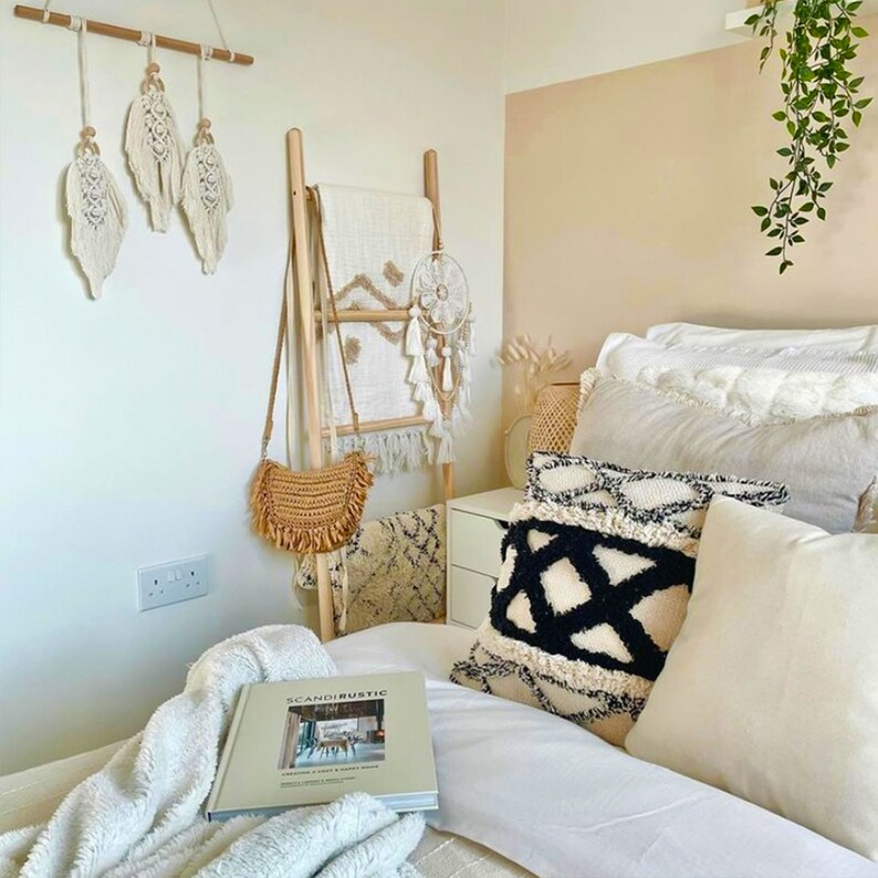 May include: A bedroom with a white and beige color scheme. The bed has a white duvet and several pillows, including one with a black and white geometric pattern. A wooden ladder leans against the wall, with a white blanket draped over it. A book titled "Scandirustic" is on the bed.