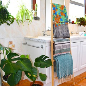 May include: A wooden ladder style towel rack with four towels hanging on it. The towels are in various patterns and colors, including blue, green, gray, and white. The rack is in a bathroom setting with a white sink and countertop.