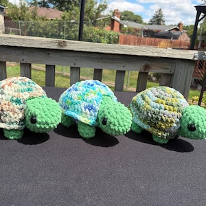 May include: Three crocheted turtle figurines with solid green heads and limbs. Each turtle features a distinctively patterned shell made of variegated yarn in shades of beige and green, blue and white, and olive green and grey, all with black eyes.