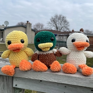 May include: Three crocheted duck plushies, one yellow, one green and brown, and one white. The plushies are sitting on a wooden railing.