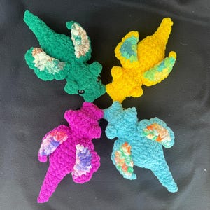May include: Four crocheted dragon plushies arranged in a cross pattern. The dragons are green, yellow, magenta, and light blue, each featuring textured bodies and wings with variegated yarn accents in shades of orange, purple, and green.