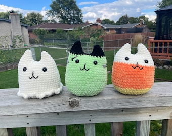 May include: Three crocheted plush toys shaped like cats, each with a different Halloween theme. The first is a white ghost, the second is a green Frankenstein, and the third is an orange, yellow, and white candy corn.