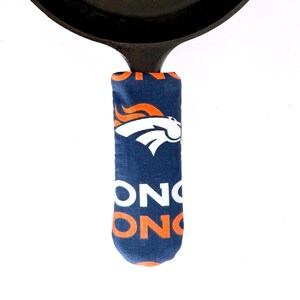 May include: A black cast iron skillet with a handle cover featuring the Denver Broncos logo in navy blue, orange, and white. The skillet has a loop for hanging and is set against a white background.