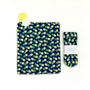 May include: A blue and green pineapple print kitchen set, including a square pot holder and a skillet handle cover. The handle cover has a white label with black text. The pineapple pattern is repeated across both items.