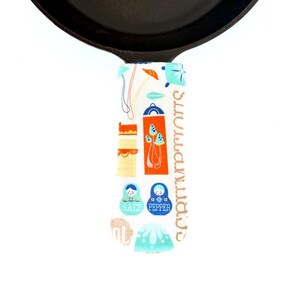 May include: A black cast iron skillet with a colorful handle cover. The handle cover features a pattern of orange, blue, and yellow illustrations of salt and pepper shakers and nesting dolls. The words "SALT" and "PEPPER" are visible.
