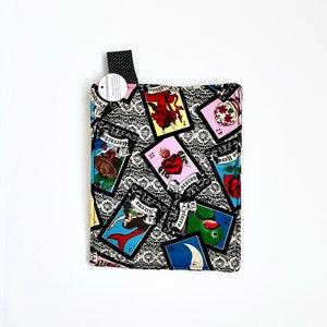 May include: A black and white pot holder with a colorful pattern of playing cards featuring images of a parrot, a mermaid, a skull, a heart, and a moon. The cards have Spanish text on them.
