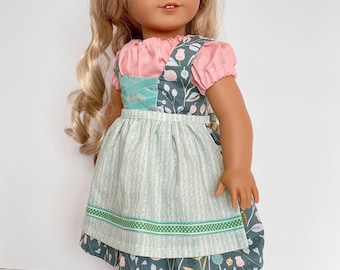 Puede incluir: Una muñeca de pelo largo rubio viste una blusa rosa de manga corta bajo un vestido de tirantes verde azulado oscuro con estampado floral. El vestido incluye un delantal a rayas verde claro con borde de lunares y zapatos verde azulado.
