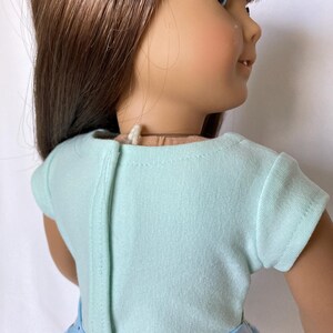 Puede incluir: Una mu&ntilde;eca con una camiseta celeste de manga corta y una falda floral azul. La camiseta tiene una costura vertical en la espalda. La falda presenta un estampado de margaritas blancas y hojas verdes sobre un fondo celeste.