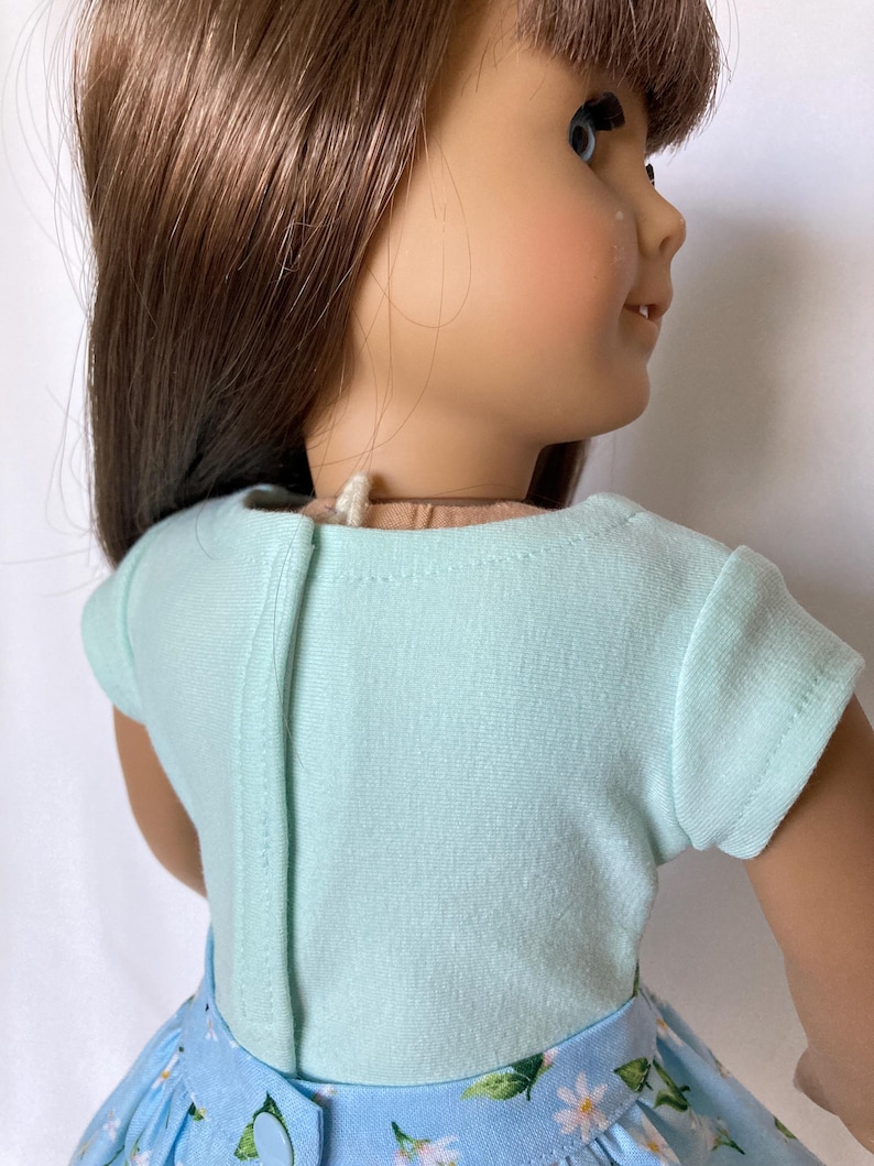 Puede incluir: Una mu&ntilde;eca con una camiseta celeste de manga corta y una falda floral azul. La camiseta tiene una costura vertical en la espalda. La falda presenta un estampado de margaritas blancas y hojas verdes sobre un fondo celeste.
