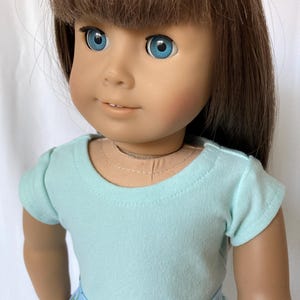 Puede incluir: Una mu&ntilde;eca con cabello casta&ntilde;o y ojos azules, vestida con una camiseta azul claro y una falda azul con un estampado floral. La mu&ntilde;eca tiene una tez clara y mira hacia la c&aacute;mara.