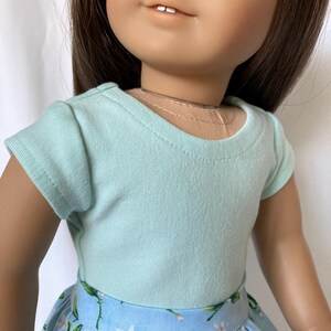 Puede incluir: Una mu&ntilde;eca con una camiseta azul claro y una falda azul con un estampado floral. La falda presenta flores blancas y hojas verdes. La mu&ntilde;eca tiene cabello casta&ntilde;o y ojos azules.
