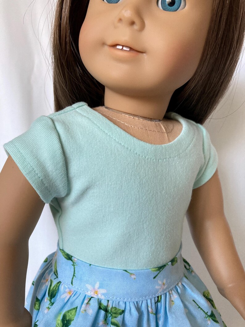 Puede incluir: Una mu&ntilde;eca con una camiseta azul claro y una falda azul con un estampado floral. La falda presenta flores blancas y hojas verdes. La mu&ntilde;eca tiene cabello casta&ntilde;o y ojos azules.