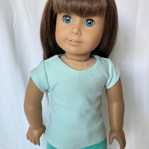 Puede incluir: Mu&ntilde;eca con cabello casta&ntilde;o y ojos azules, vestida con una camiseta azul claro y leggings verde azulado. La mu&ntilde;eca est&aacute; posada sobre un fondo blanco. El atuendo de la mu&ntilde;eca es sencillo e informal.