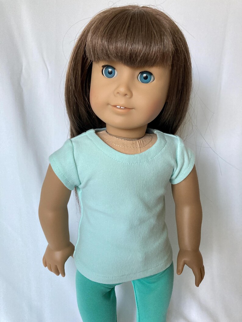 Puede incluir: Mu&ntilde;eca con cabello casta&ntilde;o y ojos azules, vestida con una camiseta azul claro y leggings verde azulado. La mu&ntilde;eca est&aacute; posada sobre un fondo blanco. El atuendo de la mu&ntilde;eca es sencillo e informal.