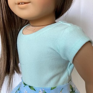 Puede incluir: Una mu&ntilde;eca con una camiseta azul claro y una falda azul con estampado floral. La falda presenta un patr&oacute;n de flores blancas y hojas verdes. La mu&ntilde;eca tiene cabello casta&ntilde;o y ojos azules.