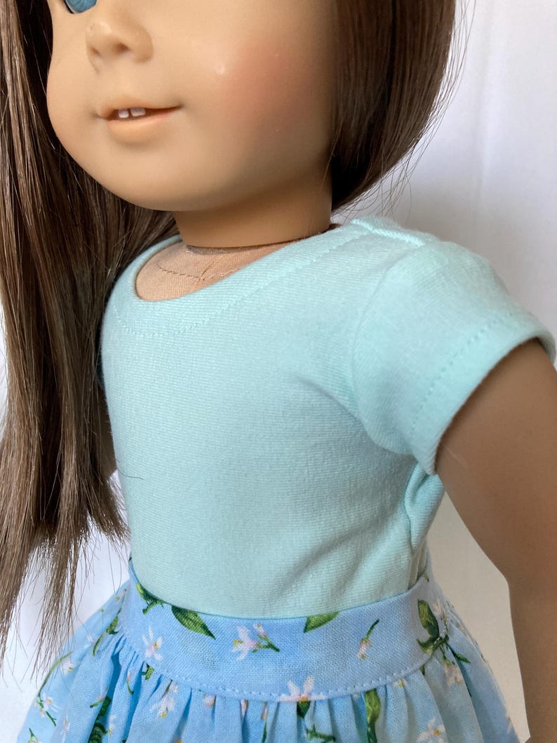 Puede incluir: Una mu&ntilde;eca con una camiseta azul claro y una falda azul con estampado floral. La falda presenta un patr&oacute;n de flores blancas y hojas verdes. La mu&ntilde;eca tiene cabello casta&ntilde;o y ojos azules.