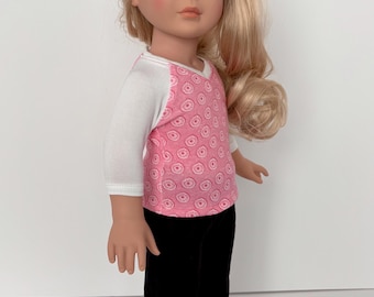 Puede incluir: Una muñeca de cabello rubio largo y ojos azules que viste una camiseta raglán blanca y rosa con estampado de donas y mangas tres cuartos, combinada con pantalones negros lisos y zapatos sin cordones rosas perforados.