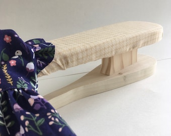 Puede incluir: Una herramienta de prensado de madera con una parte superior acolchada, cubierta con una tela beige con un patrón a cuadros. La herramienta está diseñada para presionar telas, como un vestido con estampado floral.